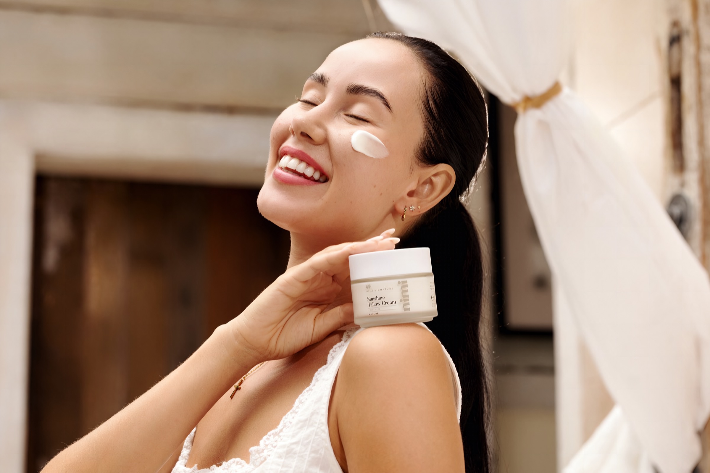 A Nimi customer laughing in warm daylight with Sunshine Tallow Cream on her cheek, holding the jar